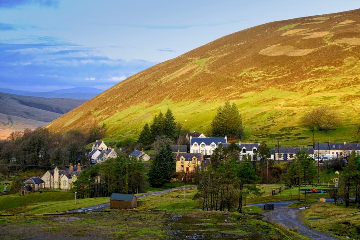 Stone cottages and white-harled houses scattered along the floor of a high valley, beneath a steep heather hillside lit gold and green by low sun, with bare moorland hills fading into the distance