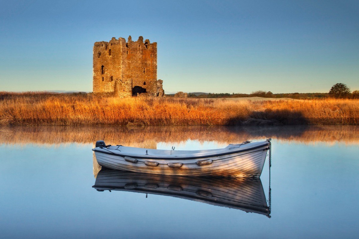 The ruined stone tower of Threave Castle on its island in the River Dee at sunrise, a white clinker-built rowing boat moored on water so still it mirrors the golden reed beds along the bank