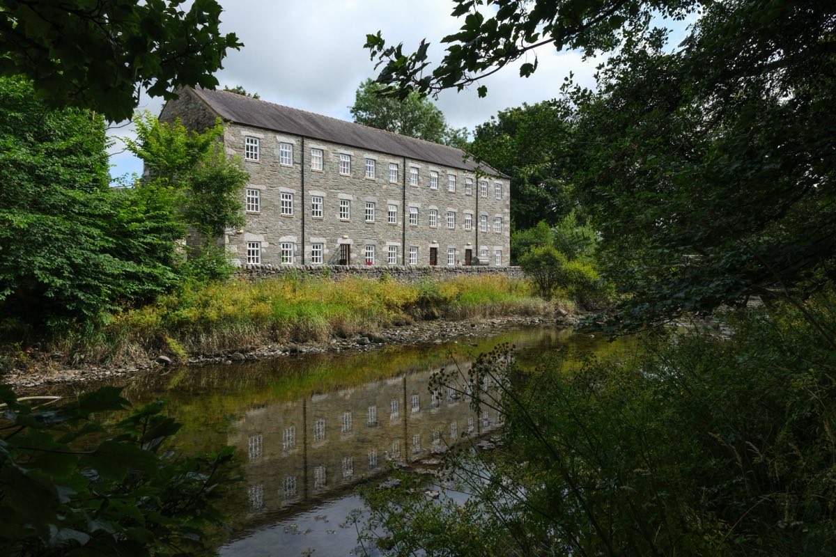The long three-storey grey stone mill at Gatehouse, rows of white sash windows under a slate roof, seen across the Water of Fleet through overhanging summer trees and reflected in the river