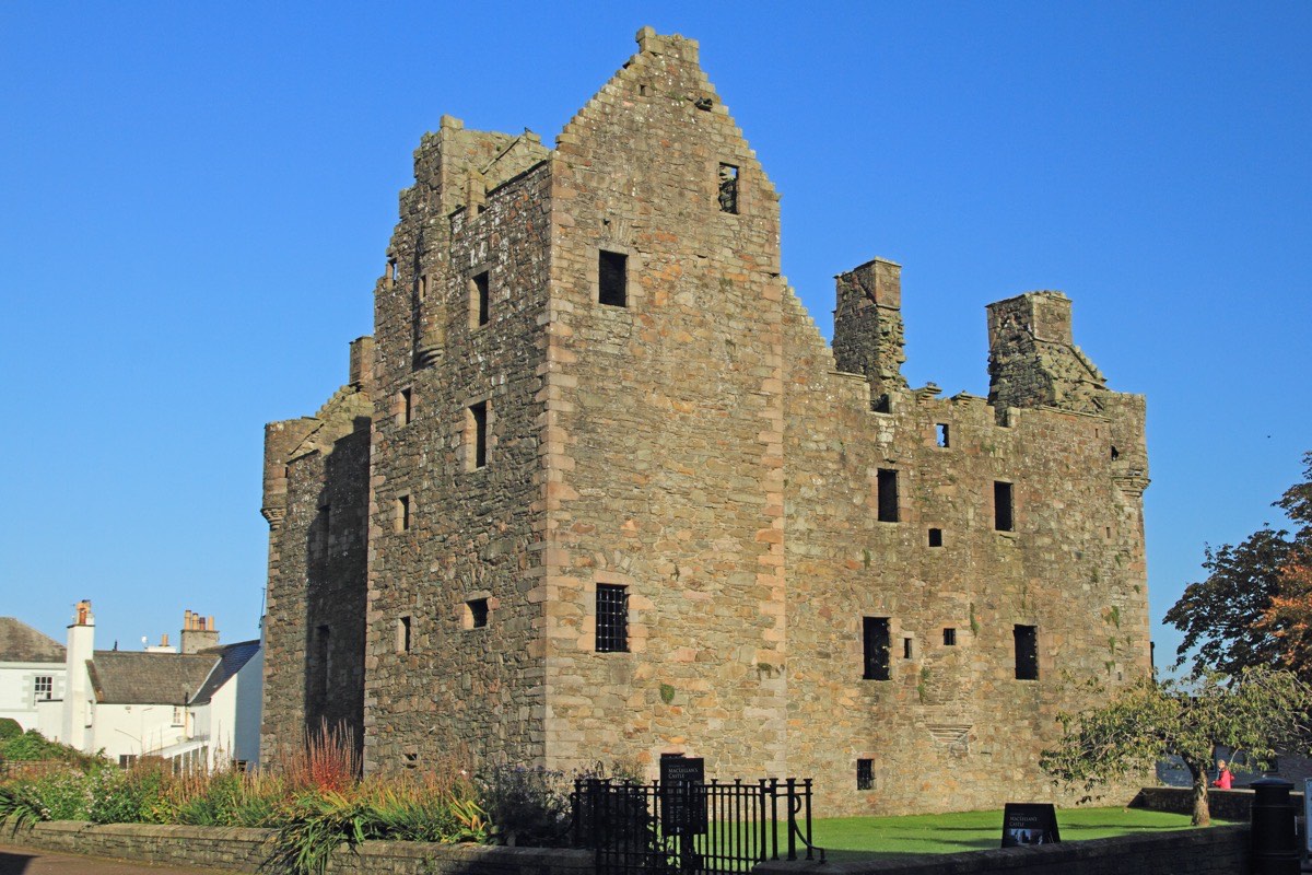 The roofless stone shell of MacLellan's Castle standing over the rooftops of whitewashed harbour houses, its empty windows and crow-stepped gables lit by low evening sun against a clear blue sky